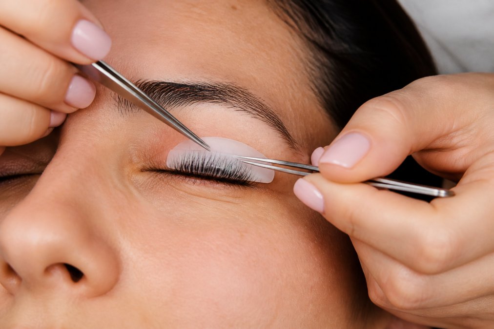 Close-up of hands using tweezers to carefully position silicone pads on an eyelid, showing proper alignment with the lash root and precise placement technique under bright clinical lighting