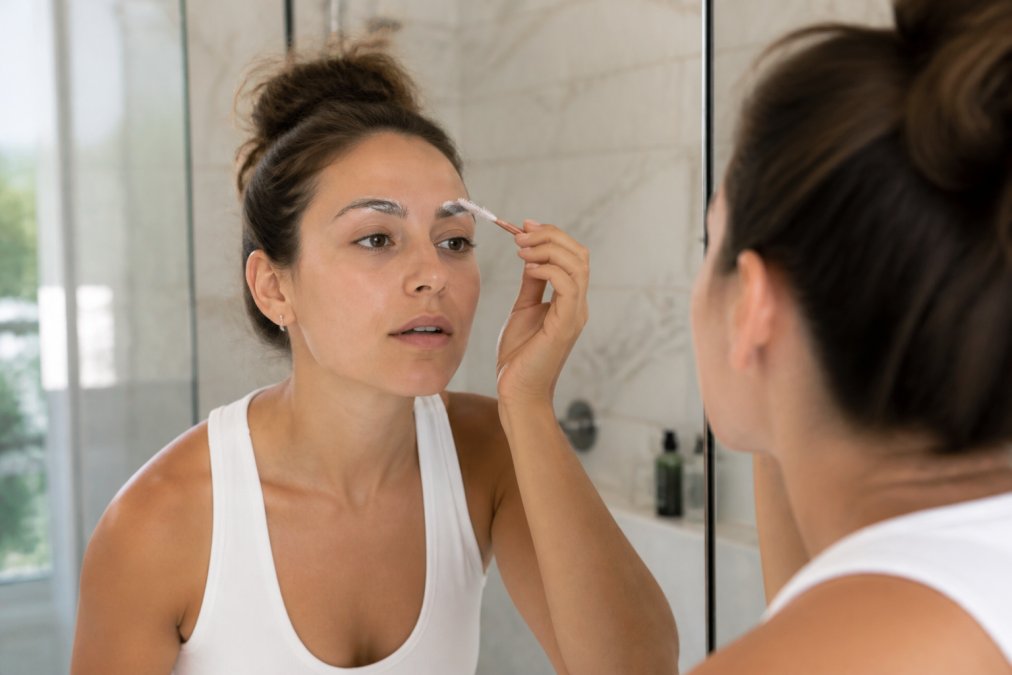 Close-up of a woman's hands applying lamination cream to her eyebrow with a spoolie brush in a bright bathroom, mirror reflection visible