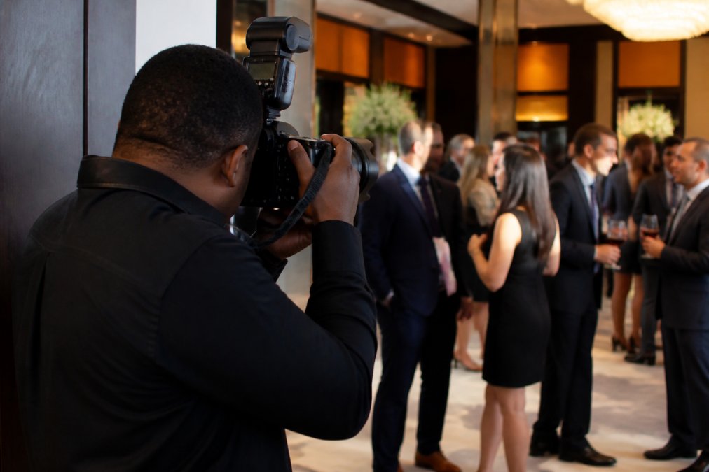 A professional photographer discreetly capturing candid moments at a corporate gala, with attendees engaged in conversation and branding elements visible in the background