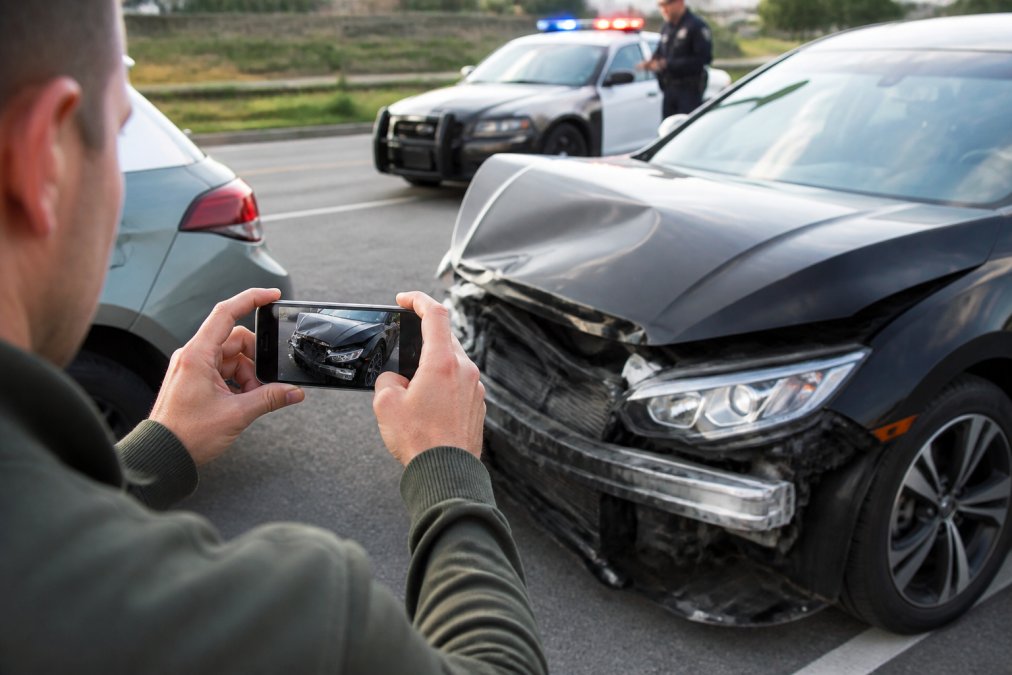 A person using a smartphone to document car accident property damage at an active scene with police nearby.