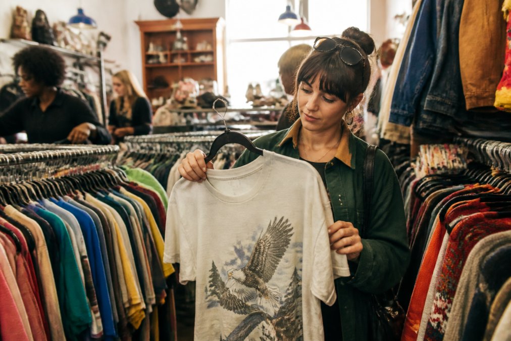 A young adult browsing racks of vintage clothing in a brightly lit thrift store, holding up a graphic tee with a thoughtful expression, surrounded by colorful second-hand garments