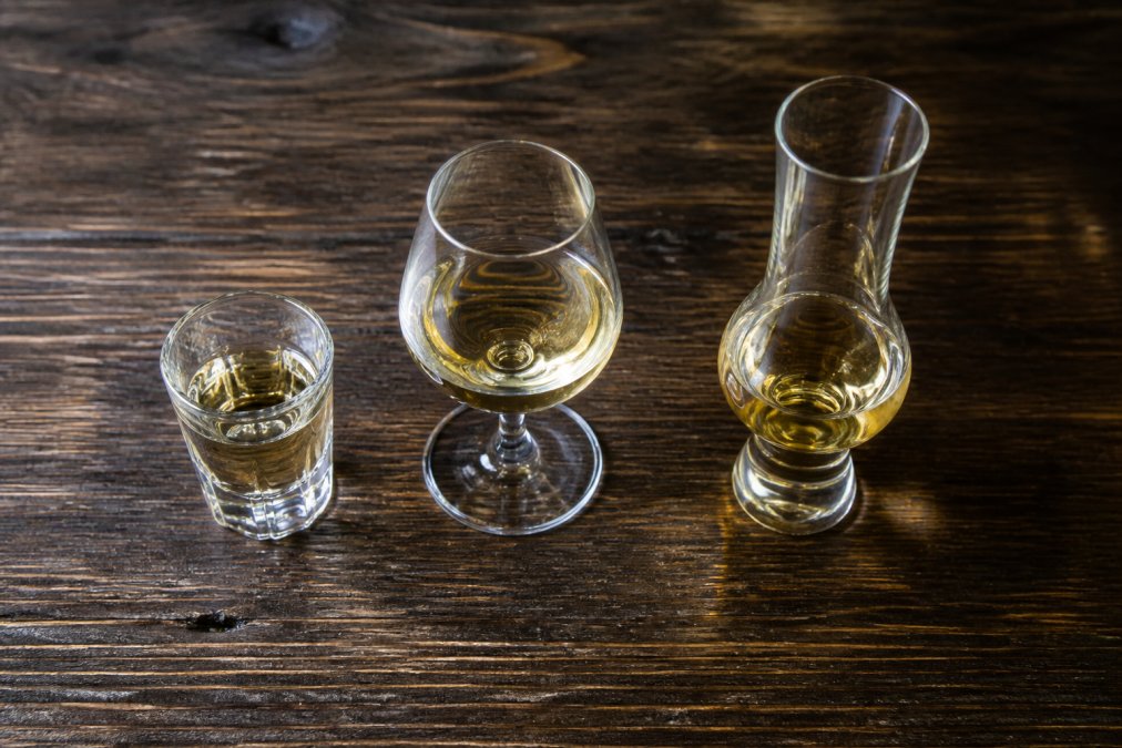 Close-up overhead shot of three different tequila glasses side by side on a dark wooden surface: a traditional shot glass, a stemmed copita glass, and a fluted nosing glass, each containing a small pour of amber-coloured reposado tequila