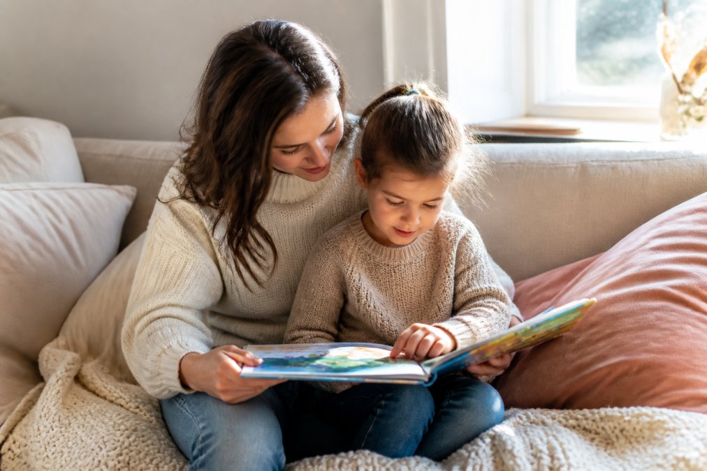 Parent and child reading together on a cozy couch with soft blankets and pillows, warm natural light from a nearby window, child engaged with a picture book