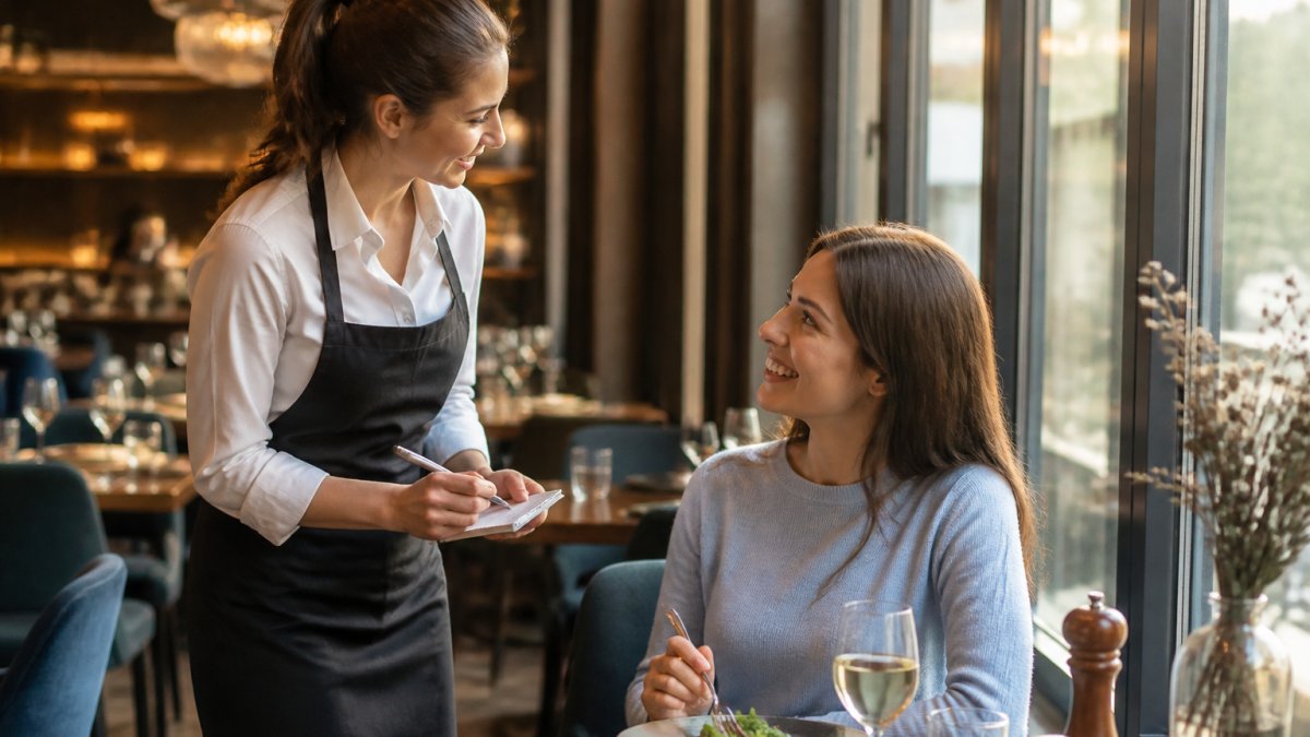 Server smiling warmly while conversing with a seated customer during their meal in a well-lit restaurant