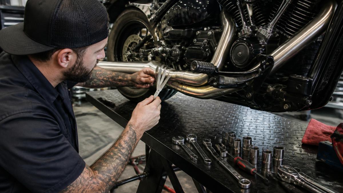 A mechanic in a workshop measuring clearance between an aftermarket exhaust pipe and a Harley-Davidson frame using a feeler gauge, with tools laid out on a workbench nearby