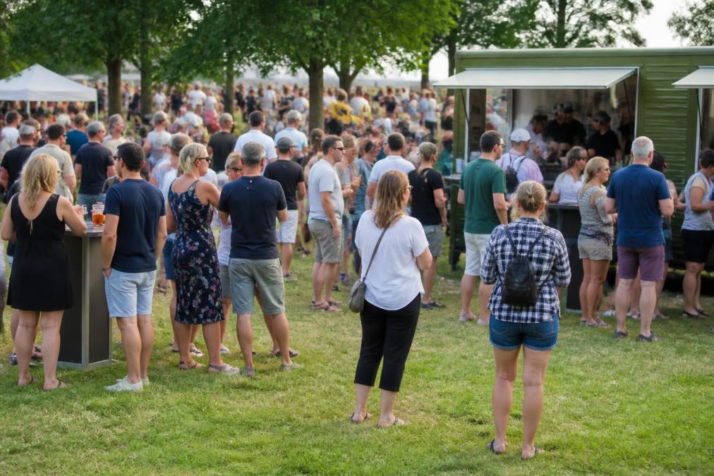 Outdoor event attendees gathered on grassy area during daytime with food and beverage service stations visible, showing typical crowd density and activity patterns