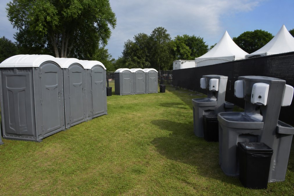 Outdoor event setup showing properly spaced portable restrooms and hand wash stations arranged on grassy area with clear signage, accessible pathways, and adequate spacing between units for service vehicle access