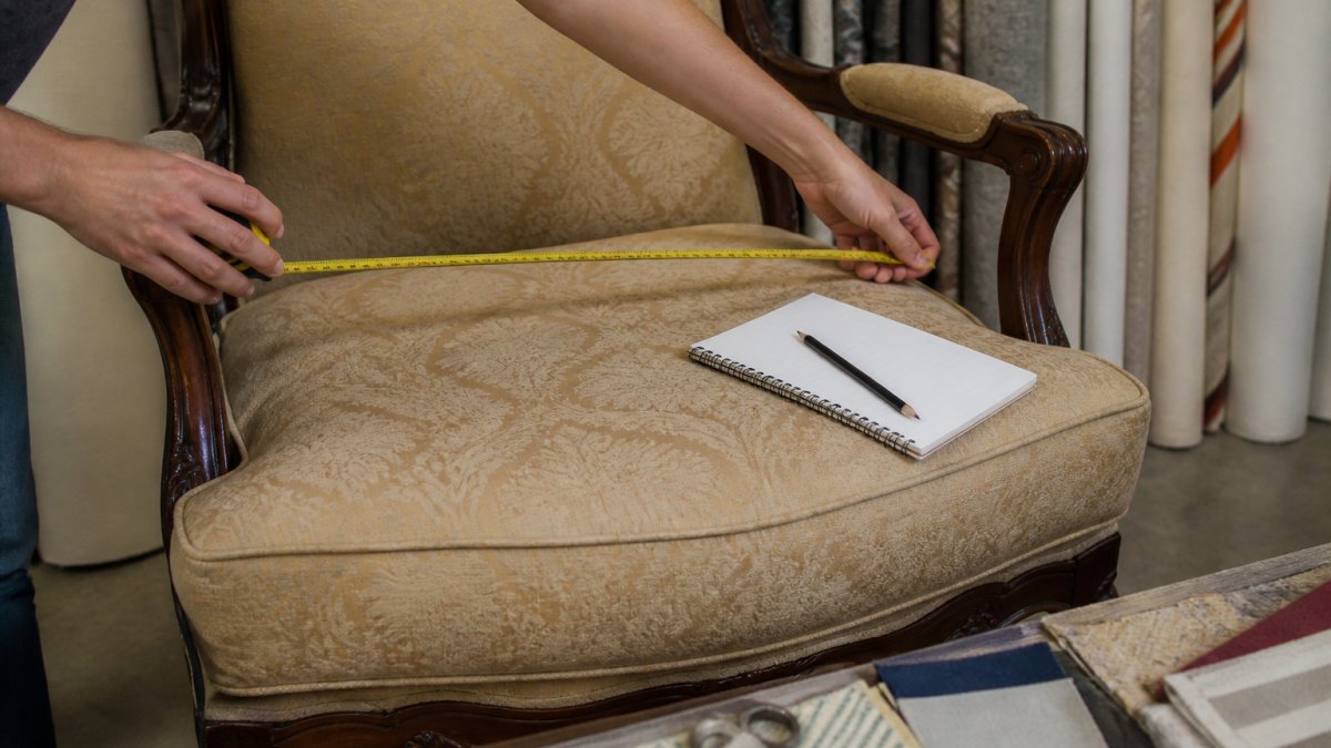 Close-up of hands holding a retractable tape measure across the seat width of a vintage armchair, with a notepad and pencil resting on the cushion beside it