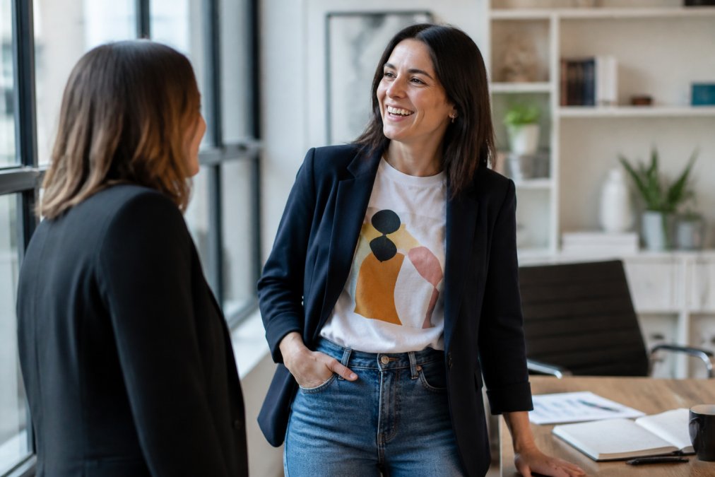 A woman in a modern office wearing a relaxed graphic tee under a structured blazer, smiling confidently while talking with a colleague near a desk, soft natural window light