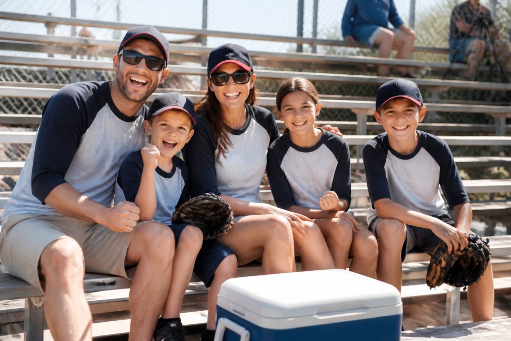 Family of five in creative fashion baseball shirts cheering on bleachers at a sunny youth game