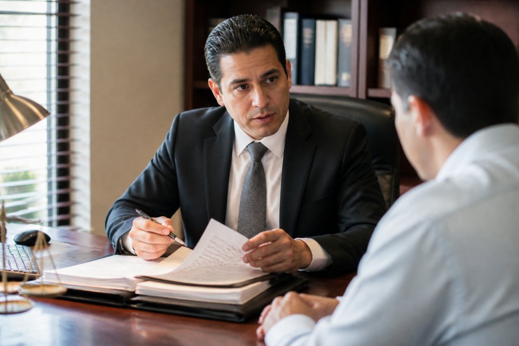 Professional attorney in business suit meeting with client across wooden desk in law office, reviewing case documents with serious focused expression