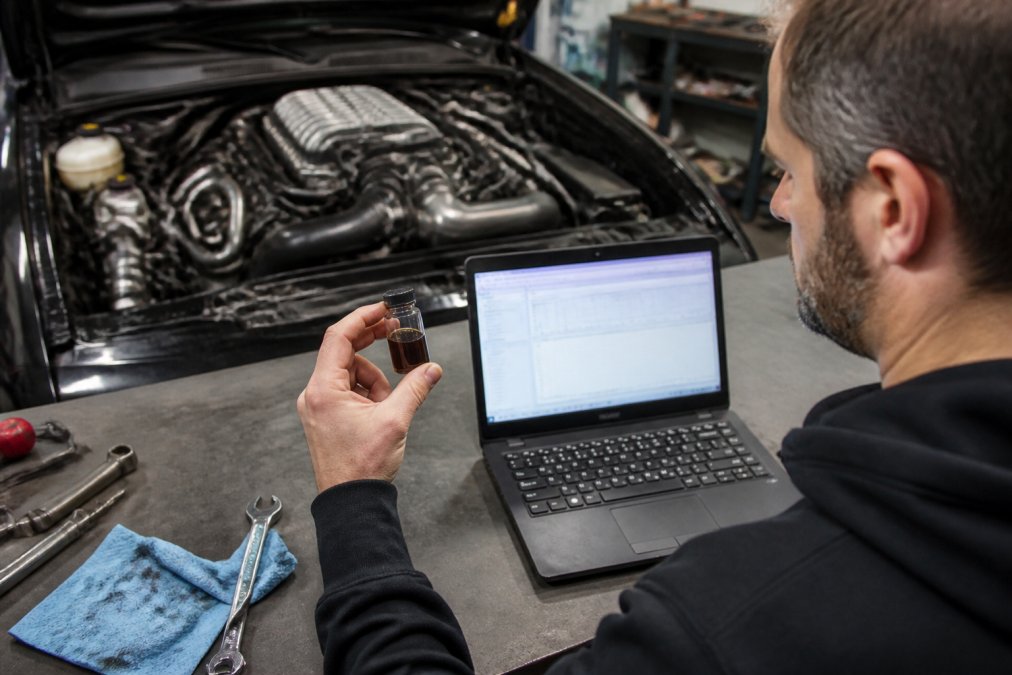 A mechanic in a home garage holding a small glass vial of used engine oil next to a laptop showing diagnostic data, with a supercharged engine bay visible in the background