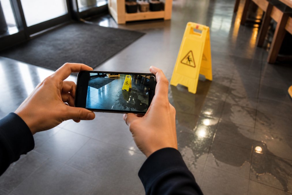 Close-up of a person's hands photographing a wet floor hazard with a smartphone, with accident scene details and wet surface visible in the background