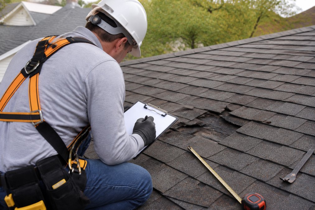 Professional roof inspector in safety gear and harness examining roof damage up close, taking detailed notes and measurements with inspection tools and clipboard visible