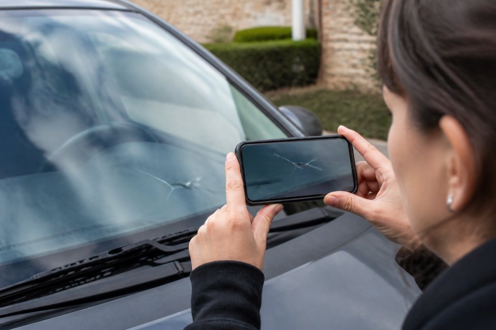 Person photographing windshield damage with smartphone in natural daylight, showing a clear crack in the glass with the phone camera focused on the damage area