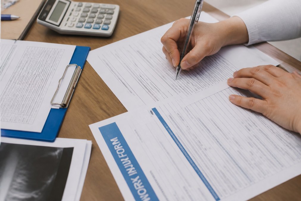 Close-up of hands filling out workers compensation forms on desk with pen, medical documents, and wage calculation sheets visible