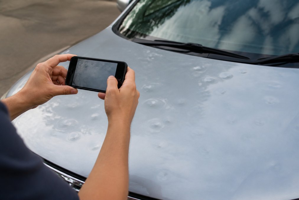 Close-up of a vehicle's hood showing multiple hail dents with a smartphone camera positioned to capture the damage details for insurance documentation
