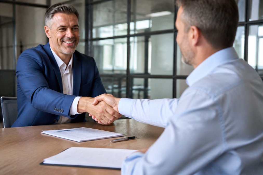 Two business professionals shaking hands across conference table with acquisition documents and pen, natural office lighting