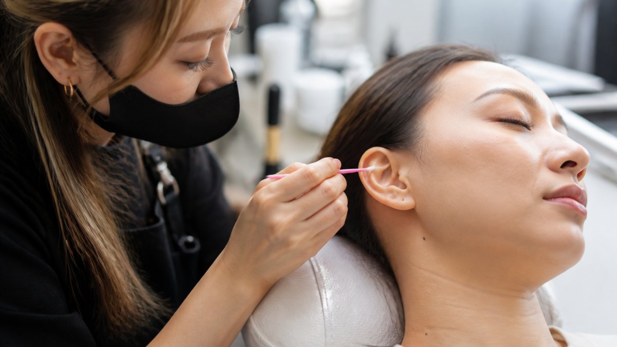 Close-up of a lash technician applying a small patch of adhesive behind a client's ear with a microbrush, client relaxed with eyes closed in a bright, clean salon