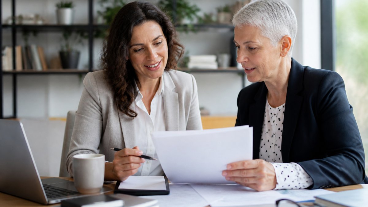 A small business owner and an HR consultant reviewing PEO contract documents and benefit plan summaries at a desk in a bright office, natural window light, papers and laptop visible