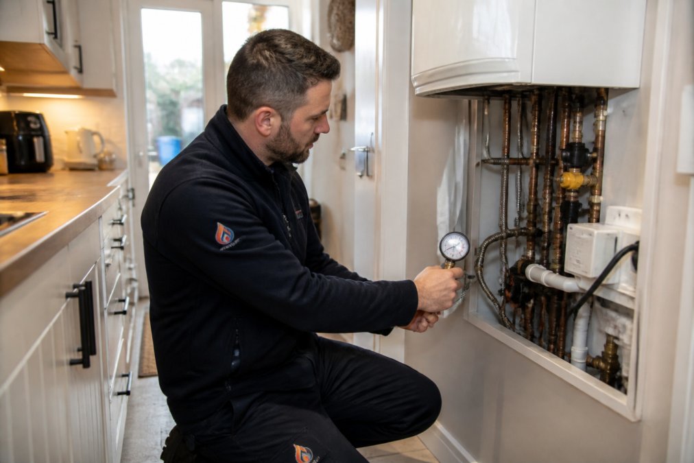 A gas engineer in branded workwear kneeling beside a modern combi boiler in a bright kitchen, holding a pressure gauge and inspecting the pipework