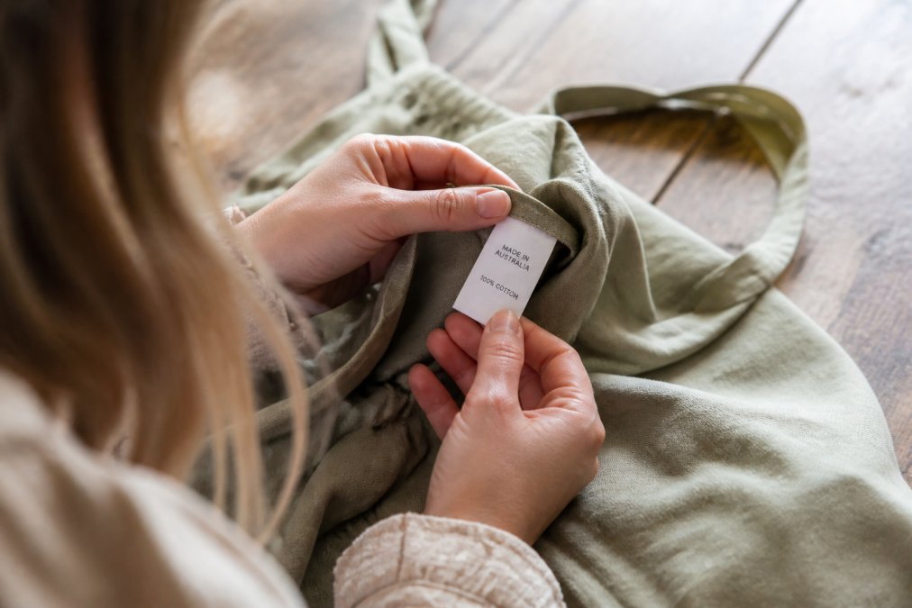 Close-up of a woman's hands turning over a garment care label inside a natural-fibre dress, soft daylight, wooden table, reading the country of origin text