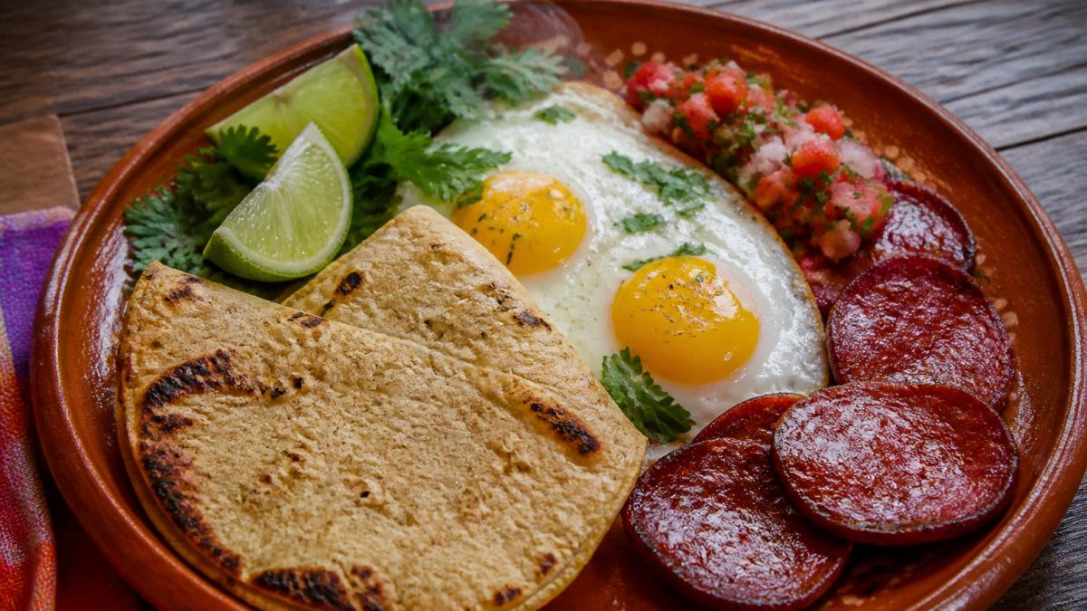 Close-up overhead shot of fresh Mexican breakfast plate with perfectly cooked eggs, chorizo slices, warm corn tortillas, fresh cilantro, lime wedges, and vibrant salsa on rustic ceramic plate with steam rising