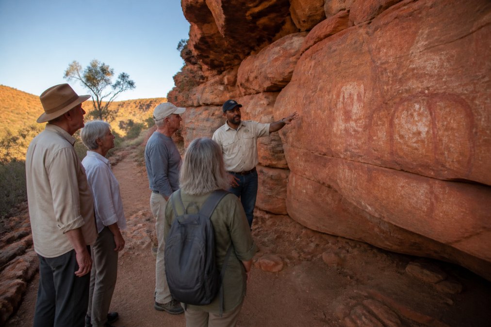 A small group of mature travellers walking with an Aboriginal guide along a red-rock landscape trail in outback Australia, the guide gesturing toward ancient rock art on a sandstone wall, warm afternoon light casting long shadows across the ochre landscape