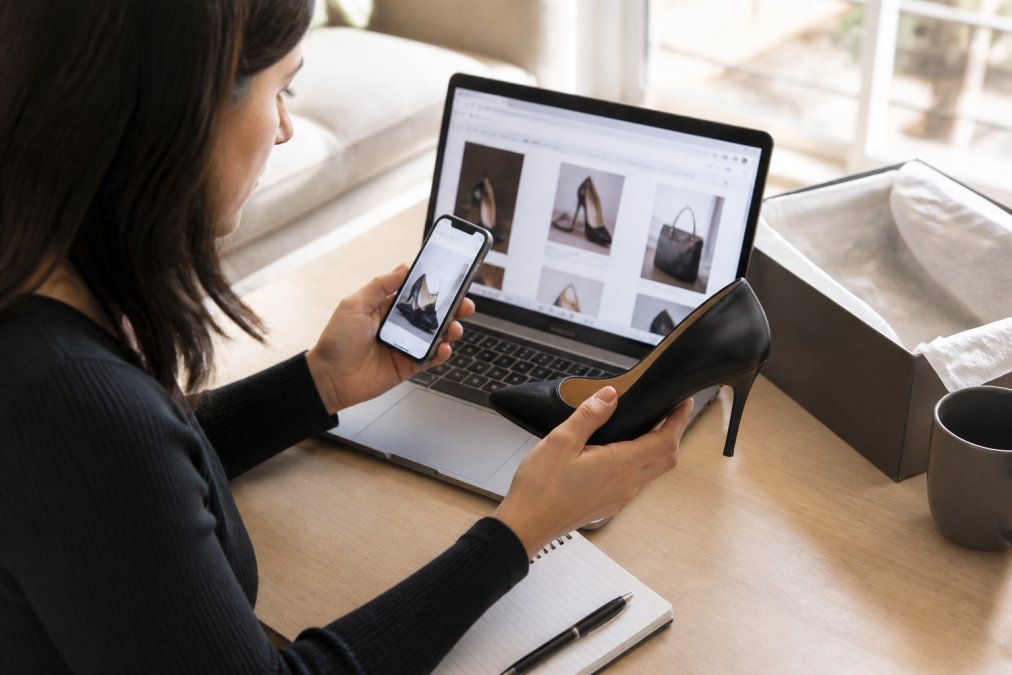 Woman at a clean desk with a laptop, smartphone, and designer shoe, carefully comparing product photos with seller information while researching brand authenticity online