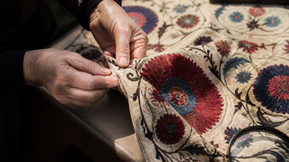 Close-up of hands examining suzani textile embroidery stitches and fabric details under natural daylight, highlighting thread quality and hand-embroidered patterns
