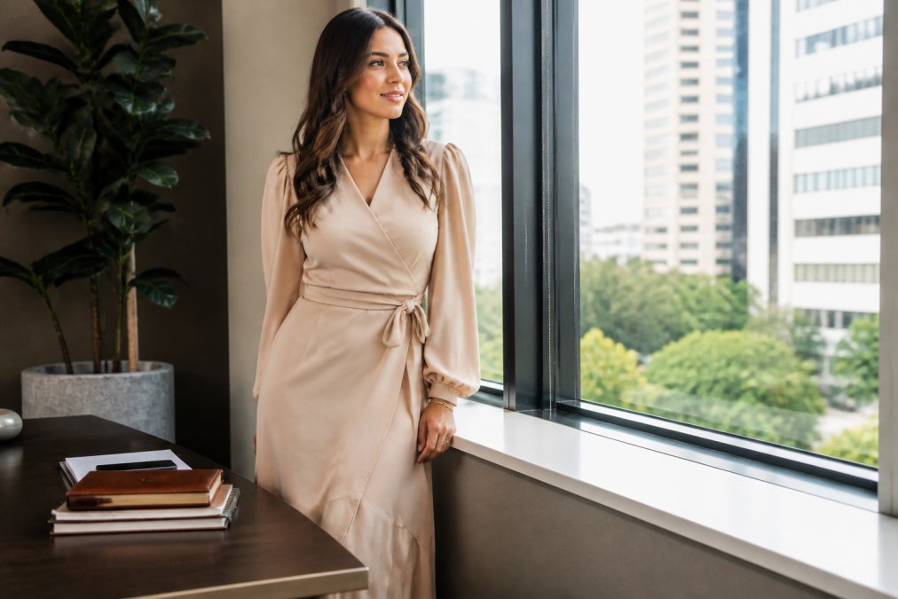 Woman wearing a stylish boutique dress in a professional office setting with natural window lighting, demonstrating how curated pieces work for everyday professional wear