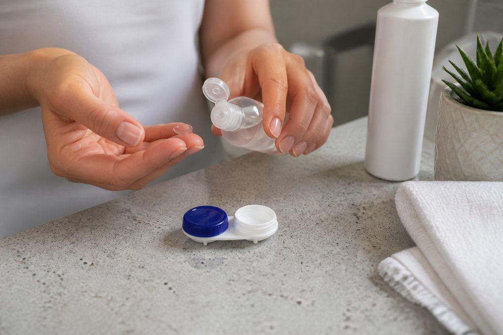 Close-up of hands properly cleaning and handling contact lenses with solution and lens case on a bathroom counter with natural lighting