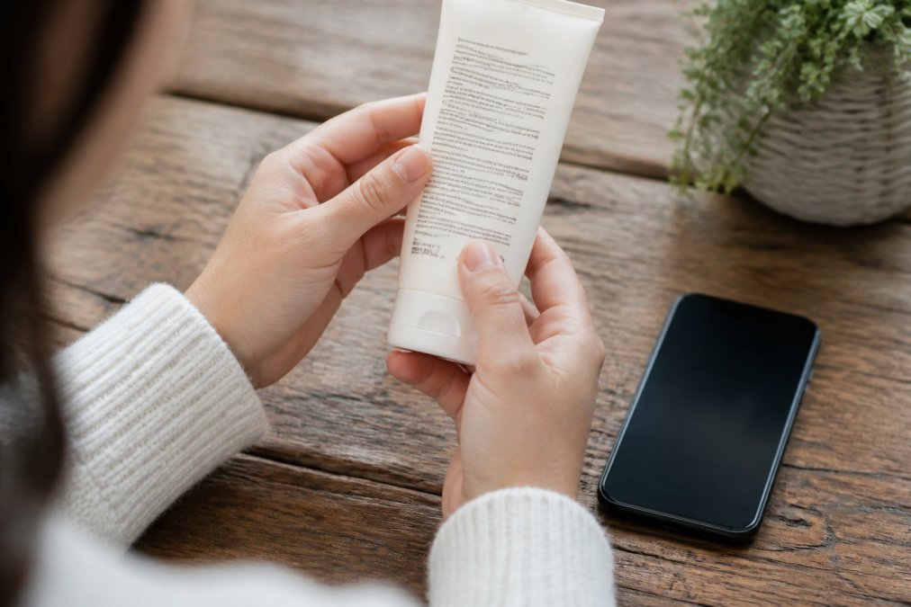Close-up of a person's hands holding a beauty product while reading the label and packaging in natural daylight, with a smartphone nearby showing a cruelty-free verification website