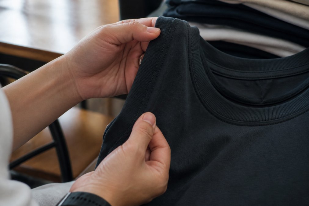 Close-up of hands examining a folded graphic tee, inspecting double-needle stitching along the side seam and collar ribbing detail with natural daylight