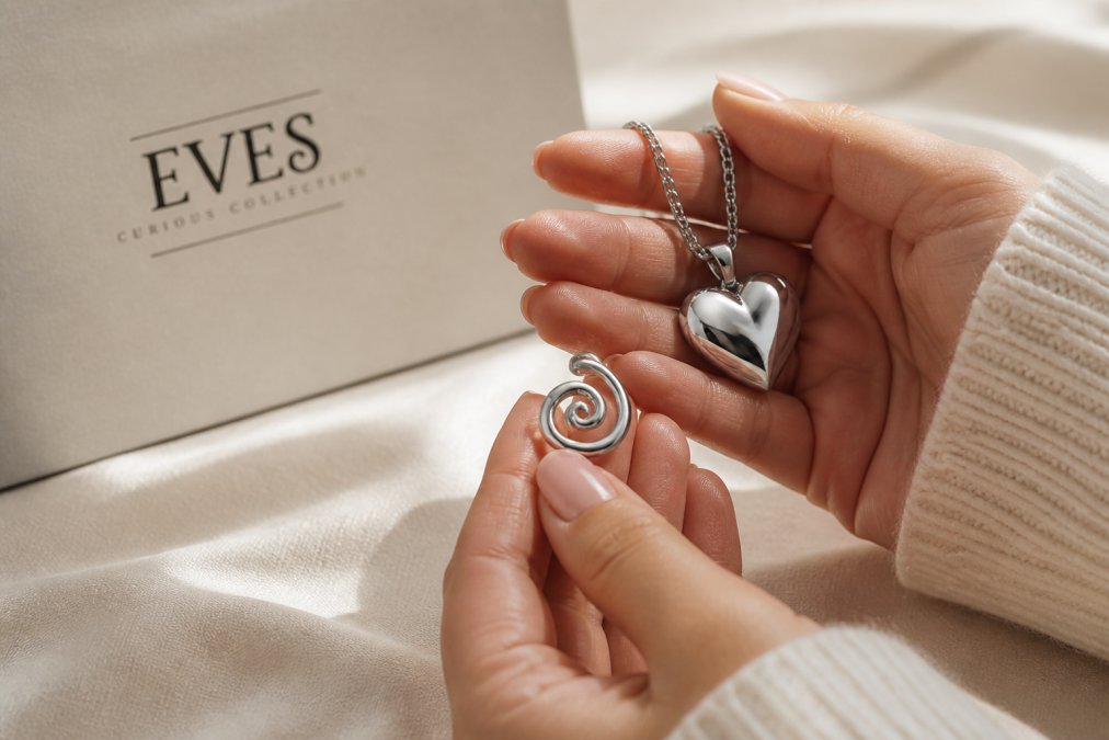 Close-up of hands holding a stainless steel heart pendant and a silver spiral ring on a soft fabric surface, natural light highlighting the metal finish