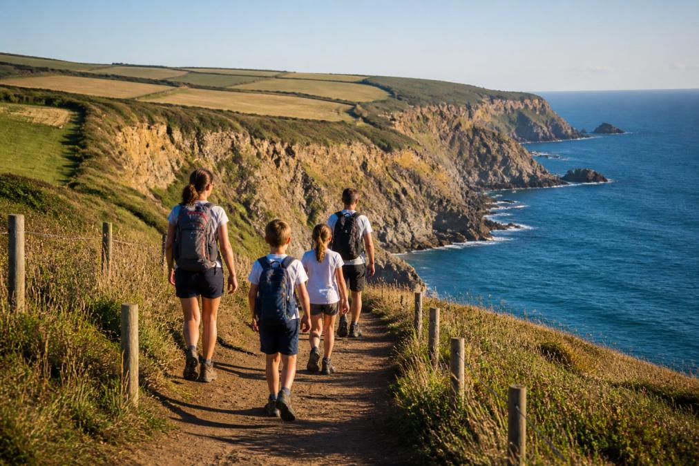Family walking along a dramatic coastal cliff path with golden cliffs and blue sea below, sunlight casting long shadows across the trail