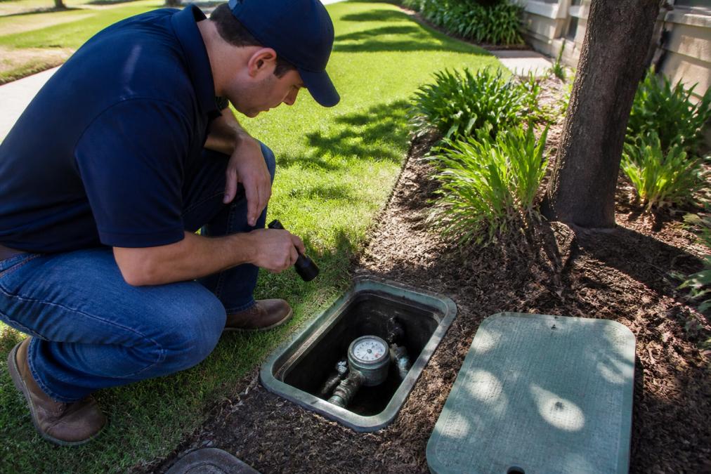 A homeowner crouching in front of an outdoor water meter box, reading the dial with a flashlight, residential yard setting, daylight