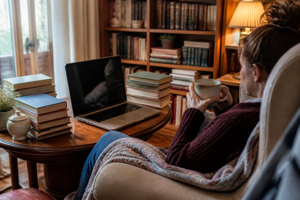 Person sitting in comfortable reading chair with a cup of tea, surrounded by stacks of independent and rare books on nearby table, laptop open showing book discovery platform, soft warm lighting from lamp