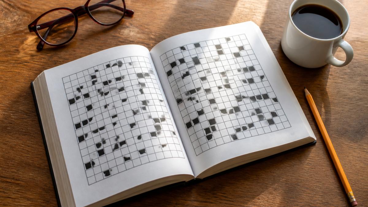 Overhead shot of an open large-print crossword puzzle book on a wooden table beside a sharpened pencil, a cup of coffee, and a pair of reading glasses, with soft natural window light