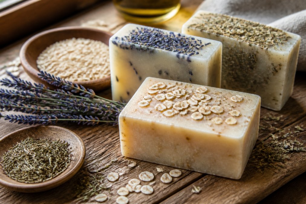 Close-up of artisanal soap bars with visible natural ingredients like oatmeal, lavender buds, and herbs on a rustic wooden surface with warm natural window lighting