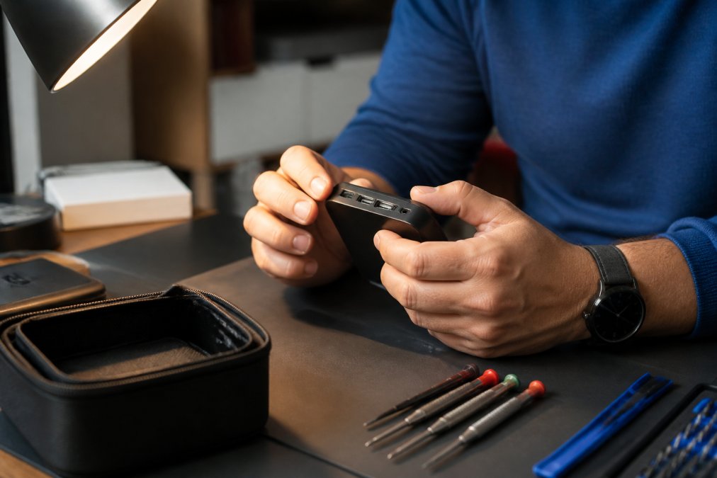 Close-up of a person's hands inspecting the seams and ports of a small electronic device under a bright desk lamp, checking for gaps and build quality