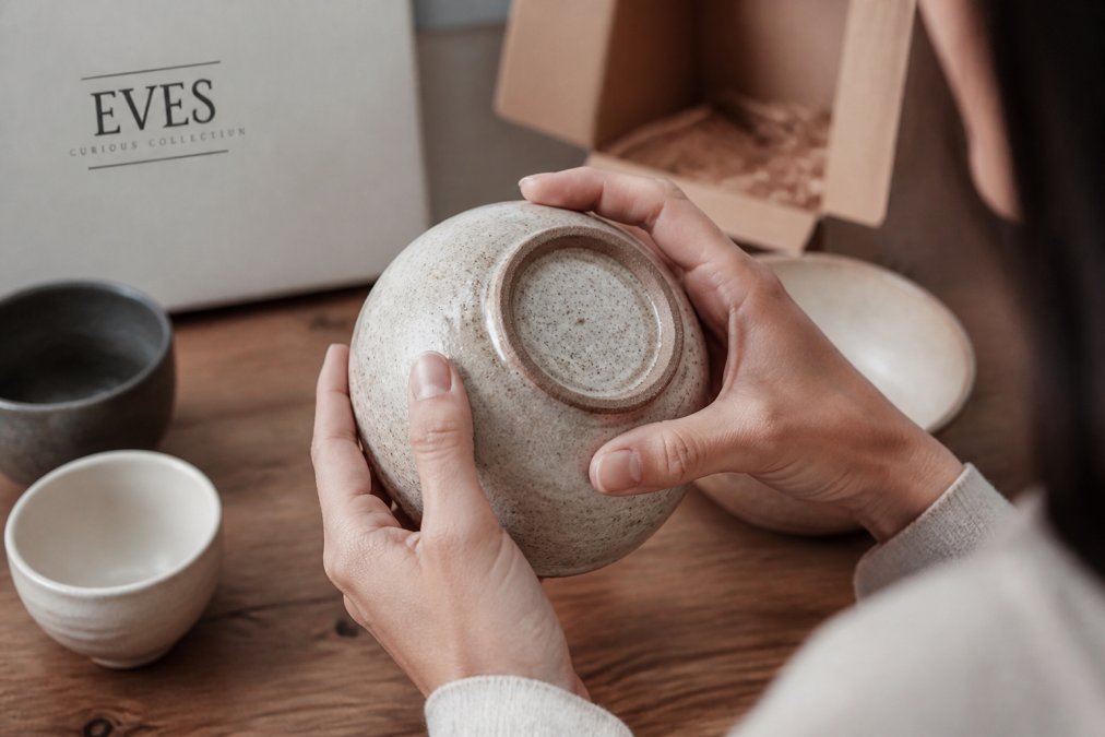 Close-up of hands examining a handcrafted ceramic bowl, turning it over to inspect the maker's mark and glaze finish on a wooden table