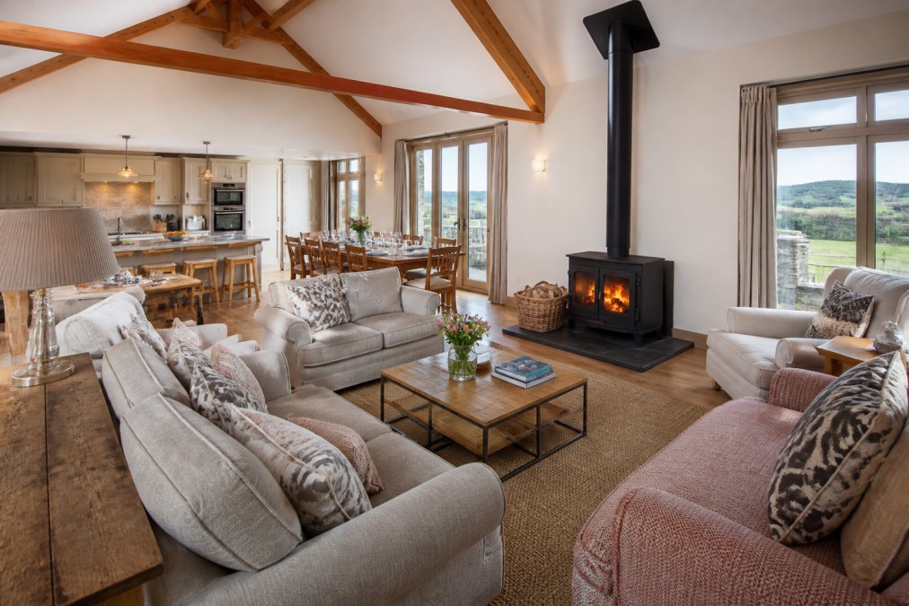 Spacious, bright open-plan living area with comfortable seating arranged around a fireplace, large dining table visible, modern kitchen in background, and countryside views through large windows