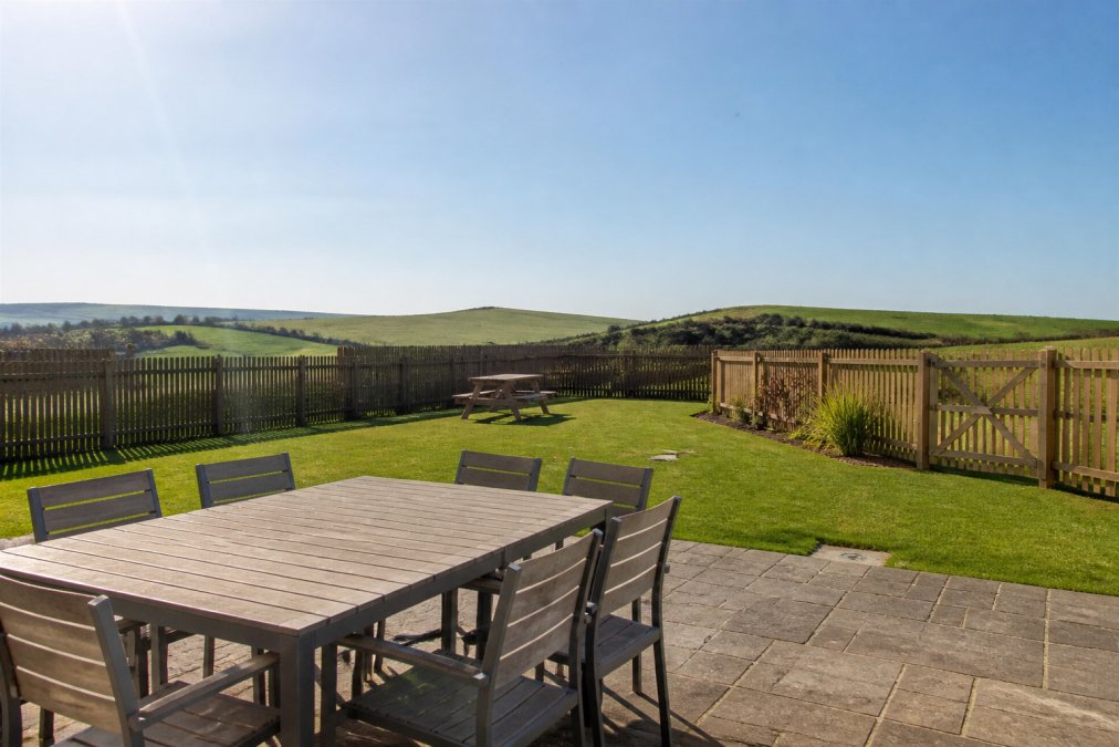 Secure enclosed garden area with sturdy fencing, manicured lawn, garden furniture, and views across countryside towards distant hills on a sunny day