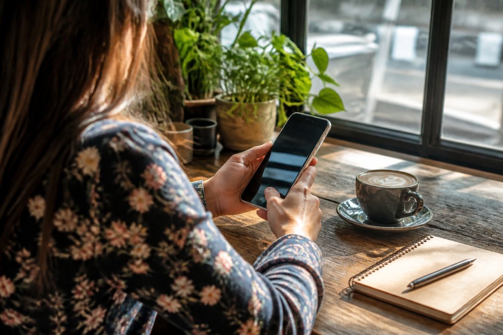 Person holding a smartphone with event discovery app open, sitting in a café with a coffee cup and notebook nearby, natural afternoon light streaming through windows