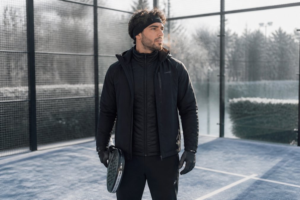 Professional padel player wearing complete cold-weather layering system (thermal base layer, dark mid-layer insulation jacket, and wind-resistant outer shell) standing on outdoor court with visible breath and frost on court surface