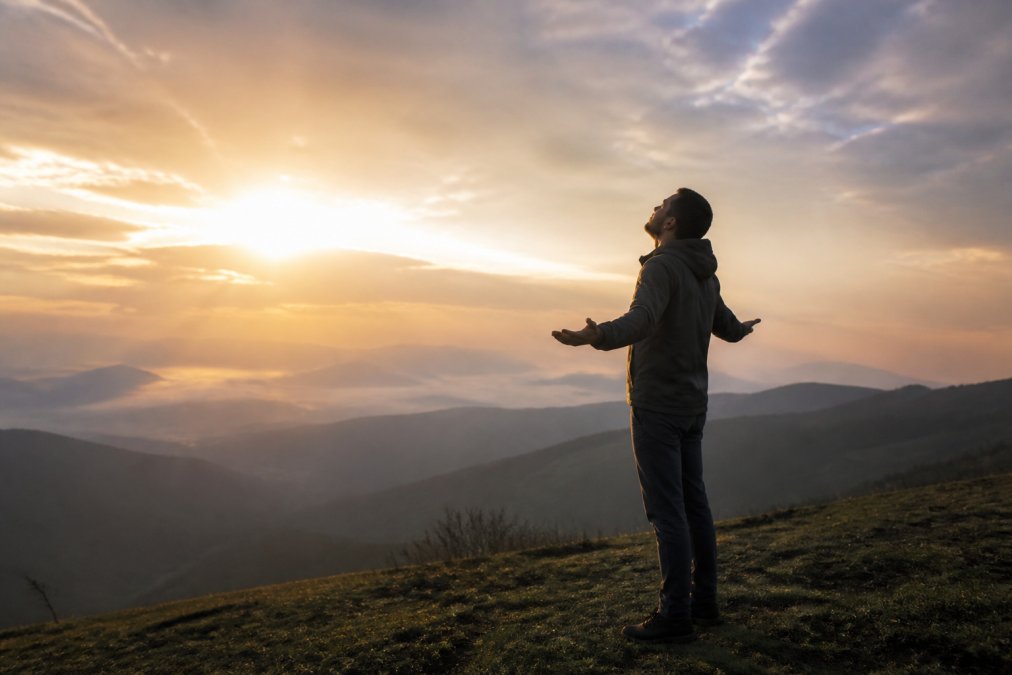 A person standing alone on a peaceful hillside at sunrise, arms slightly open, looking upward with a calm and hopeful expression
