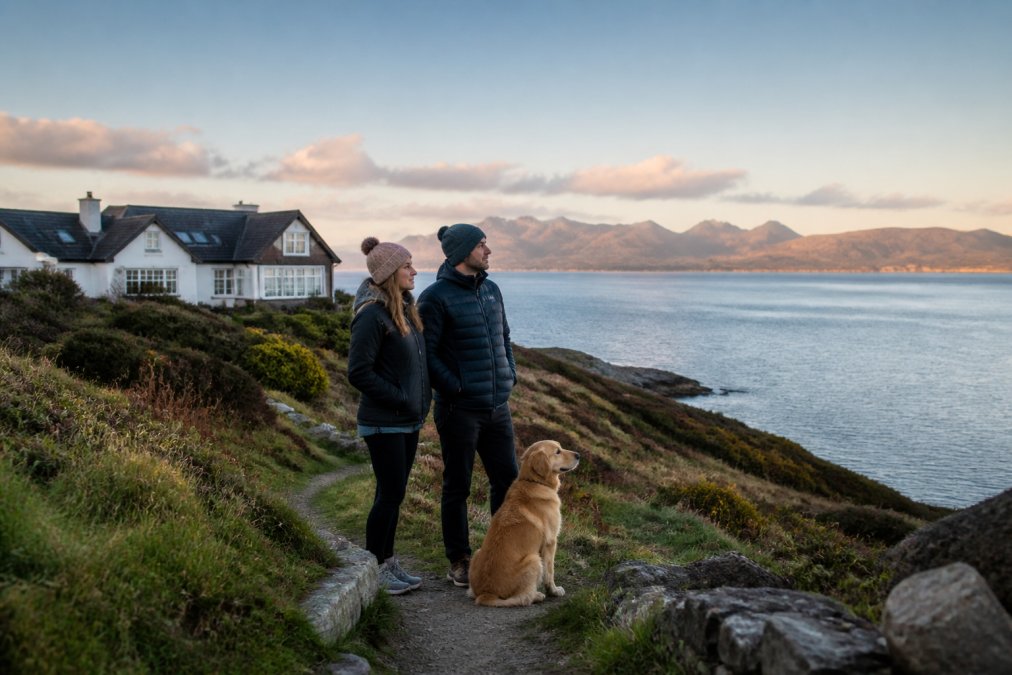 A couple standing with their golden retriever on a coastal path overlooking Loch Dunvegan, the lodge visible behind them and the Cuillin mountains on the horizon in soft evening light