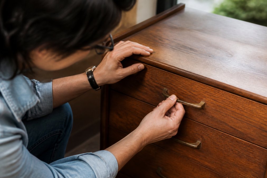 Close-up of a person's hands inspecting the wood grain and brass hardware of a vintage mid-century modern dresser, looking for signs of restoration and wear in natural window light