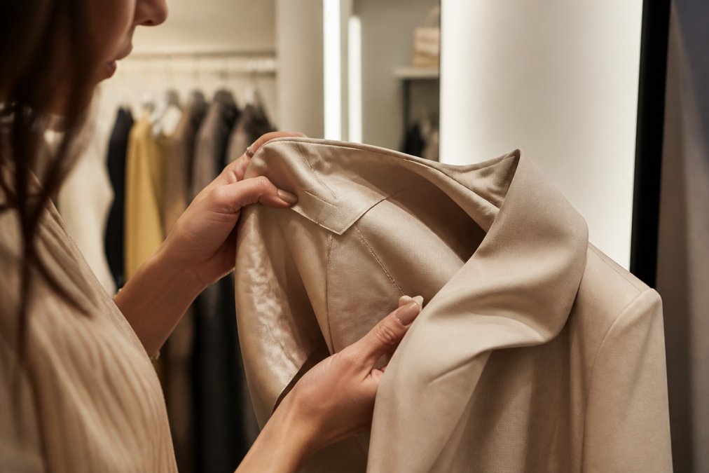 Close-up of a woman's hands examining the fabric composition label and inner seam of a neutral-toned blazer in a well-lit boutique fitting room